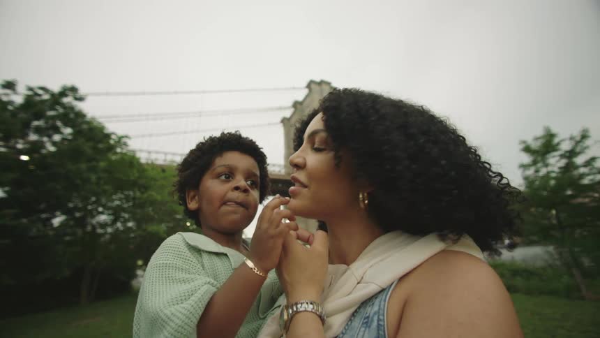 A smiling woman holds a young boy who playfully touches her face as they share a tender moment in a park witha bridge in the background