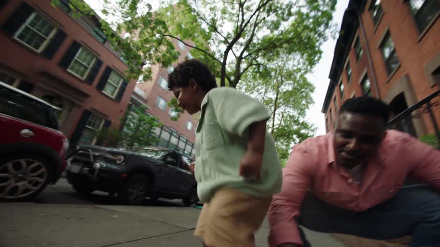A father and son draw a sun with sidewalk chalk outside their brownstone as a woman looks on from the steps
