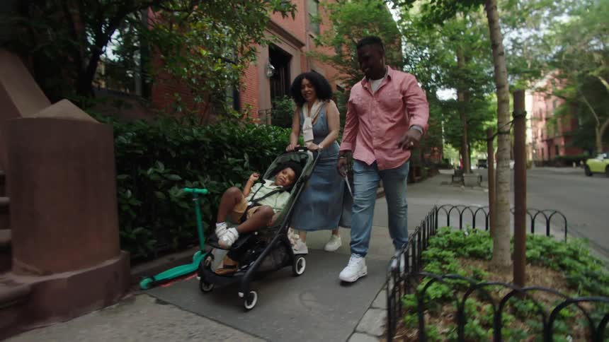 The video shows a family consisting of a father mother and child walking down a city street the child is comfortably seated in a stroller while the parents walk alongside pushing the stroller