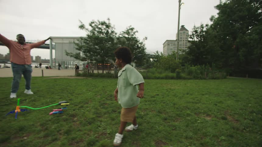 A young child in an oversized green shirt walks across a grassy park toward colorful toy equipment with an adult nearby and an urban skyline in the background