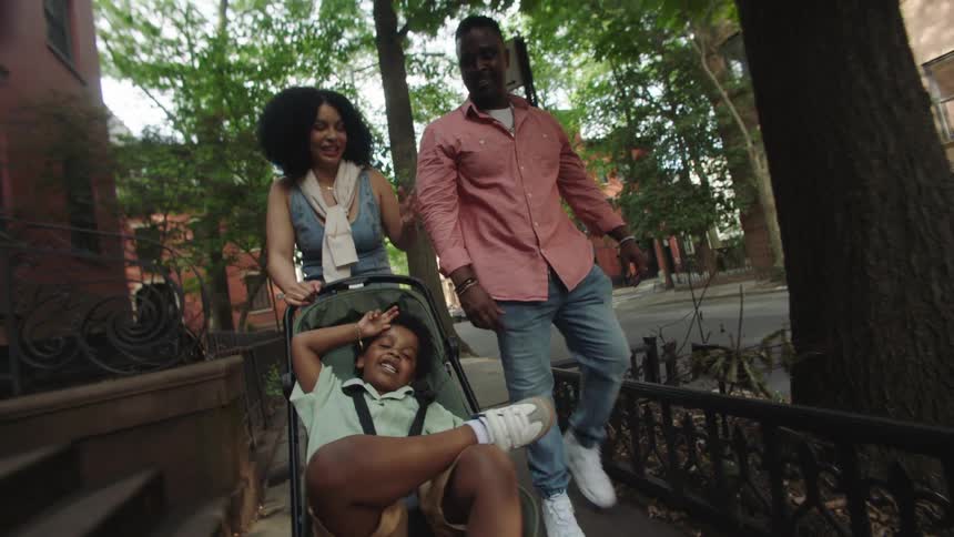 The video shows a family consisting of a woman a man and a young child in a stroller walking down a treelined sidewalk in what appears to be a residential area with brick buildings the woman is pushing the stroller while the man walks alongside and