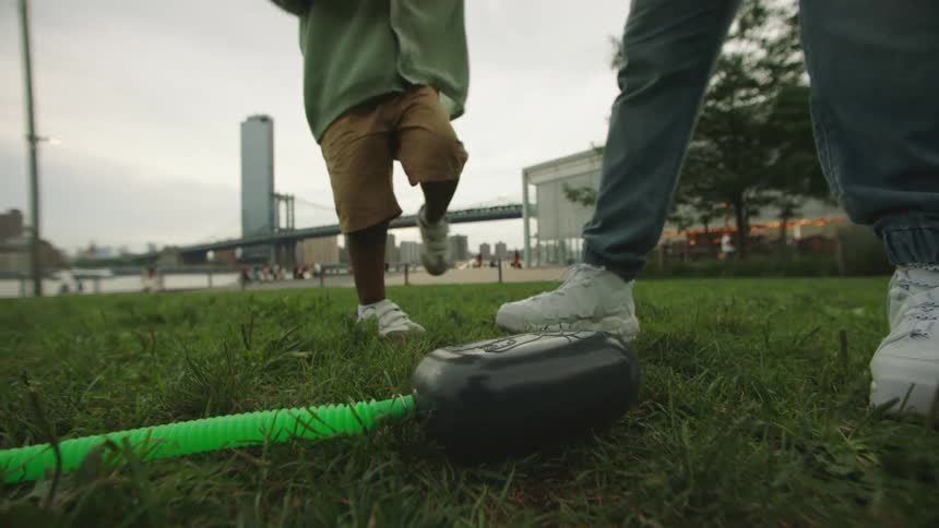 A child stomps on a black air pump attached to a green tube in a grassy park near the waterfront with an adult standing nearby and a bridge in the background