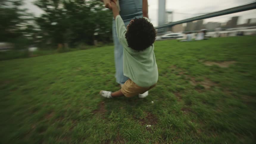 The video shows an adult and a young child possibly a mother and son playing in a grassy park area with the iconic brooklyn bridge visible in the background they are holding hands and spinning around joyfully indicating a playful interaction