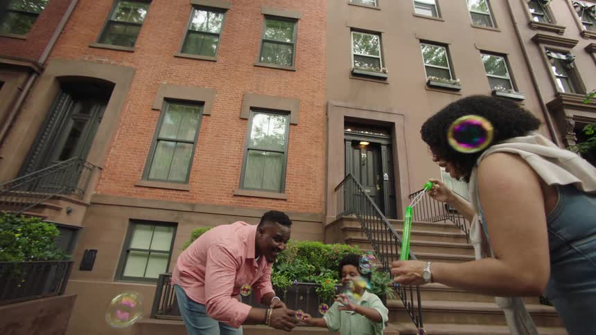 A joyful family plays with bubbles on the sidewalk in front of classic brownstone buildings with tall windows and iron railings