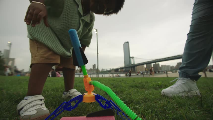 The video shows a child actively engaged in playing with a toy that launches projectiles possibly in a park setting given the grass and distant bridge the child is seen operating the toy which has green and blue components while an adults legs are vi