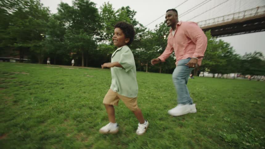 The video shows a man and a young boy running uphill on a grassy slope with the brooklyn bridge visible in the background implying they are in a park or open area near the bridge they are both smiling and appear to be enjoying themselves suggesting a p