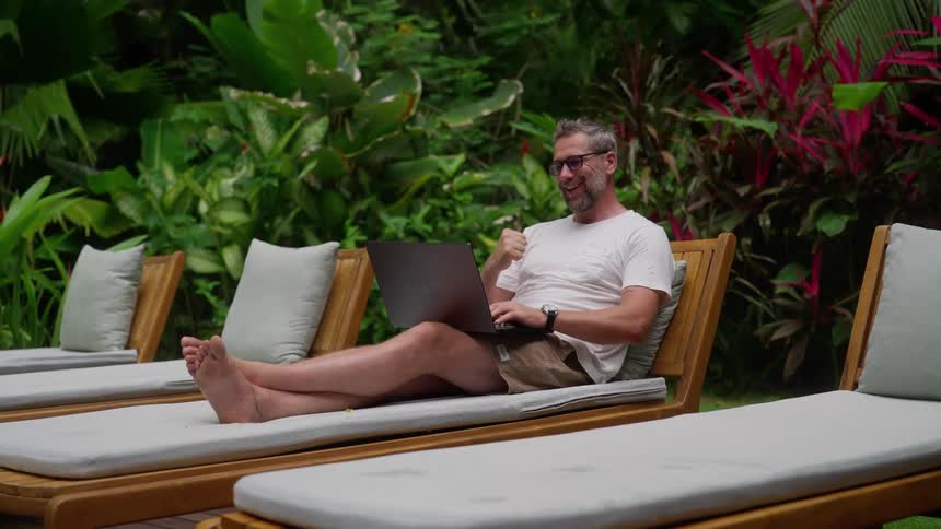 A man wearing a white tshirt sitting on an outdoor pool chair typing on a laptop and suddenly becoming excited at midday