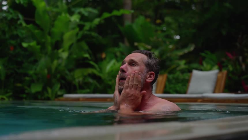 The video features a man partially submerged in a swimming pool looking out over the water the background consists of lush greenery and what appears to be outdoor lounge furniture suggesting a resort or tropical setting