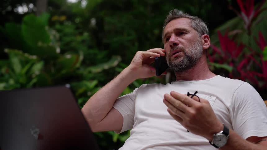 A man in a white shirt sits outdoors with a laptop engaged in a serious phone conversation surrounded by lush tropical plants