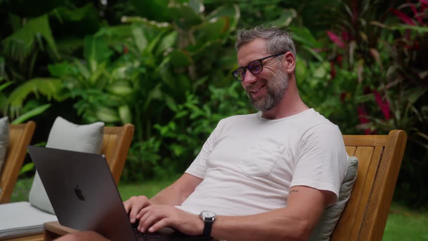 A man laughs as he uses his laptop while sitting on a chair outdoors during daytime