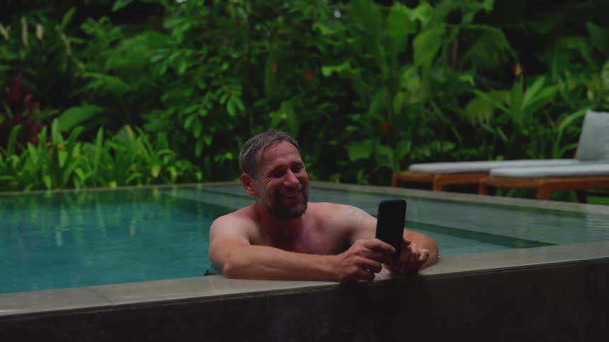 Man leans on the side of an outdoor pool during a video call on his smartphone on a summer day