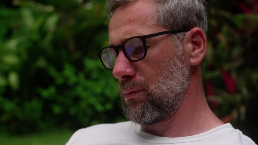 A bearded man wearing glasses and a white shirt sits outdoors in a lush garden is looking sad