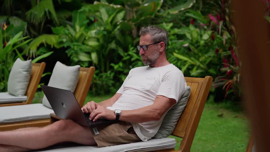 Man works on a laptop while relaxing on a lounge chair in a tropical garden