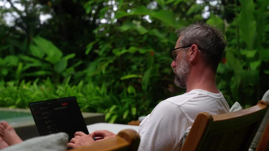 Man sitting in an environment with tropical trees typing on a laptop