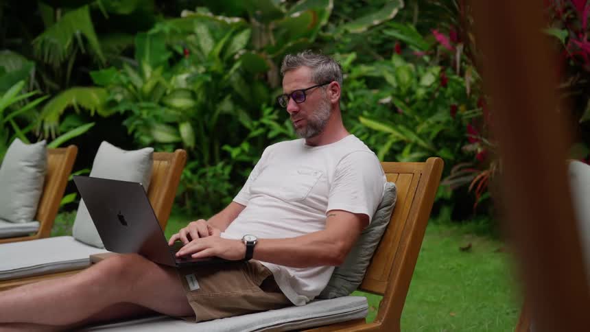 Man sitting on a sun lounger and looking at his watch and interacting with a laptop