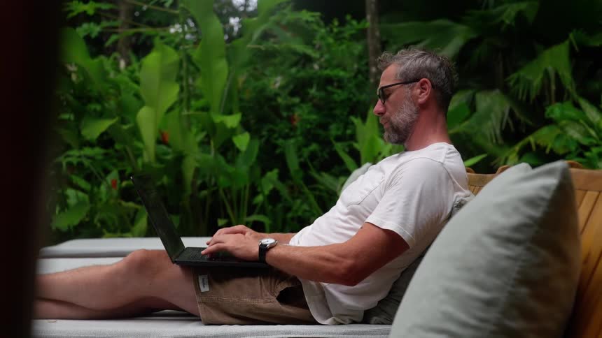 Man sitting with legs stretched out interacting with a laptop and looking at his smartwatch