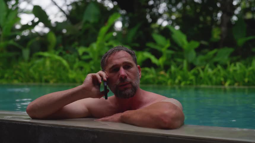 The video features a man with a beard and no shirt likely in a swimming pool or hot tub talking on a smartphone with a lush green natural background he appears relaxed and engaged in conversation