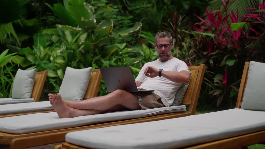 A man is typing on his laptop on a lounger outdoors