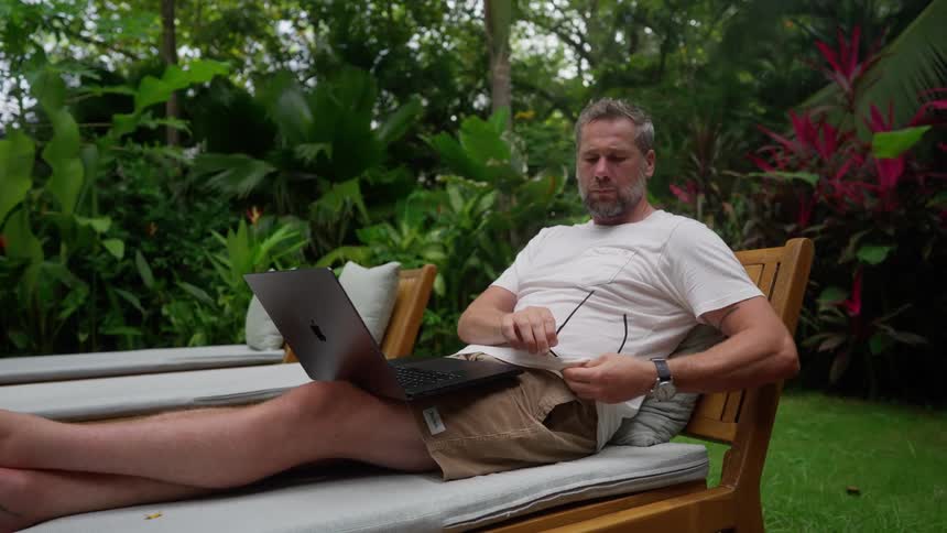 Man cleans his glasses with his shirt as he uses his laptop while sitting outdoors during daytime