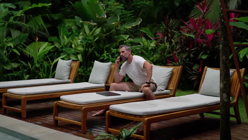 A man lounges barefoot on a cushioned chair in a lush tropical garden working on a laptop