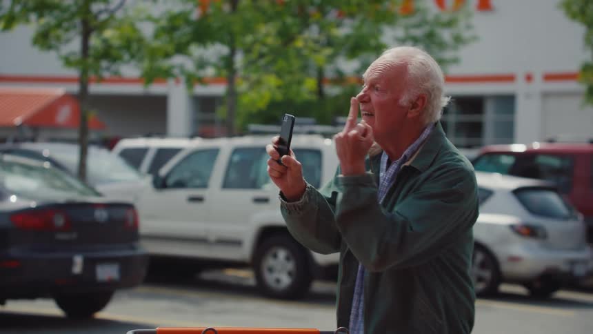 Elderly man stops while pushing a trolley in a car park to angrily make a phone recording during daytime