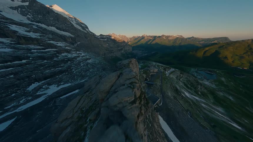 A dramatic aerial view captures a rugged mountain ridge bathed in golden light with snow patches and deep valleys stretching toward distant sunlit peaks