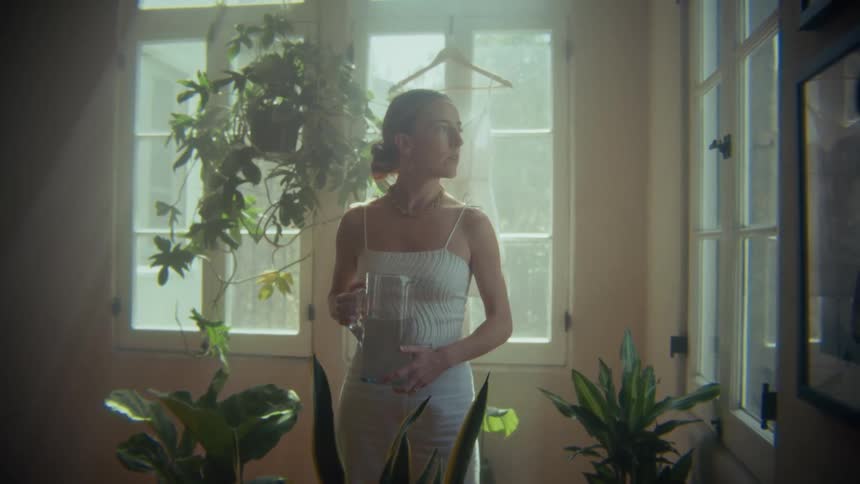 Woman stands contemplatively while carrying a glass jug in an indoor garden during daytime