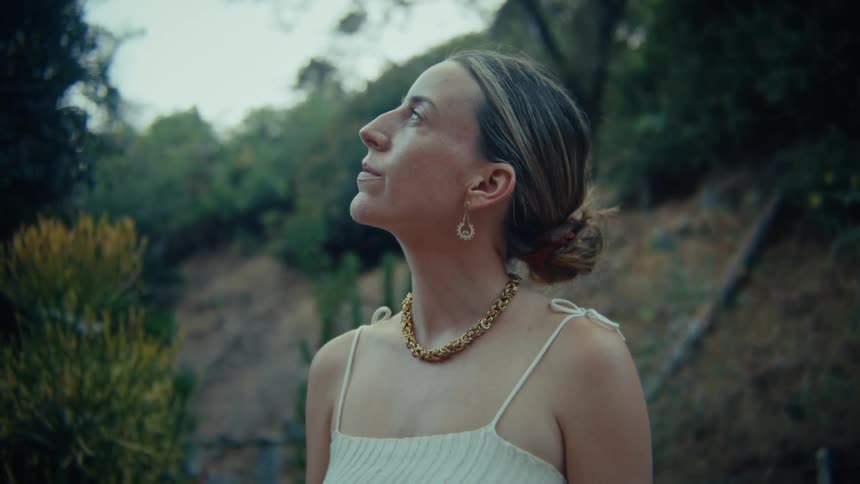 The video features a woman in profile looking upwards and slightly to the right with lush green foliage in the background suggesting an outdoor or natural setting she is wearing a lightcolored possibly white top with thin straps and a prominent gol