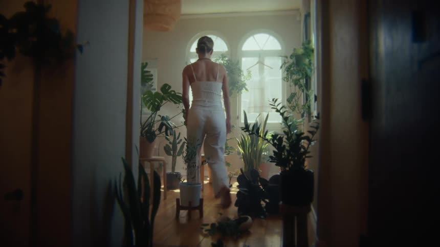A woman walks into a room filled with potted plants and then sits on a chair at midday