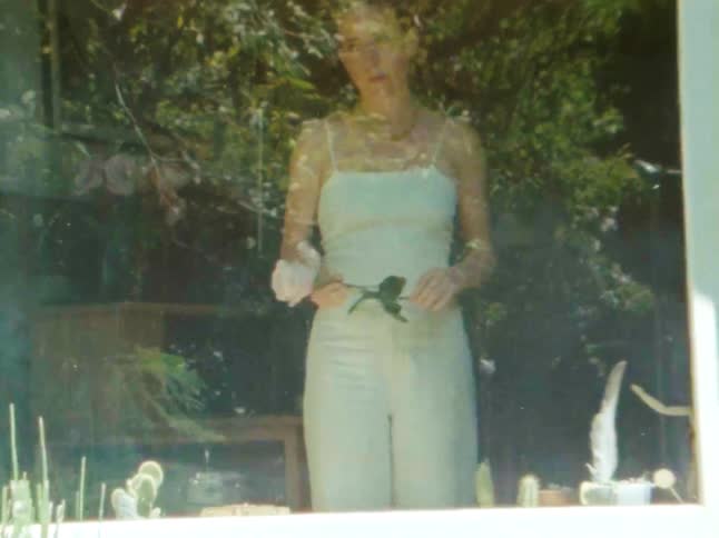 A woman in a lightcolored dress is shown in a softfocus outdoor setting gently holding and looking at a single light pink rose