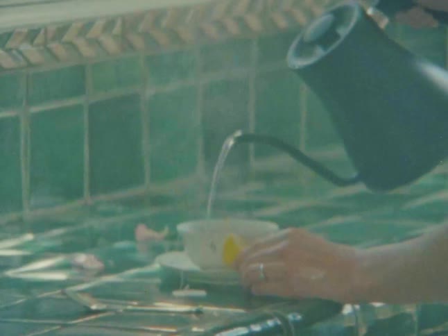 A person pours hot water from a metal kettle into a teacup on a green tiled counter with a lemon wedge resting on the saucer