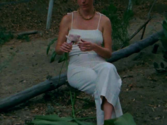 A woman in a white tank top and pants is sitting on a log outdoors surrounded by dry terrain and various plants