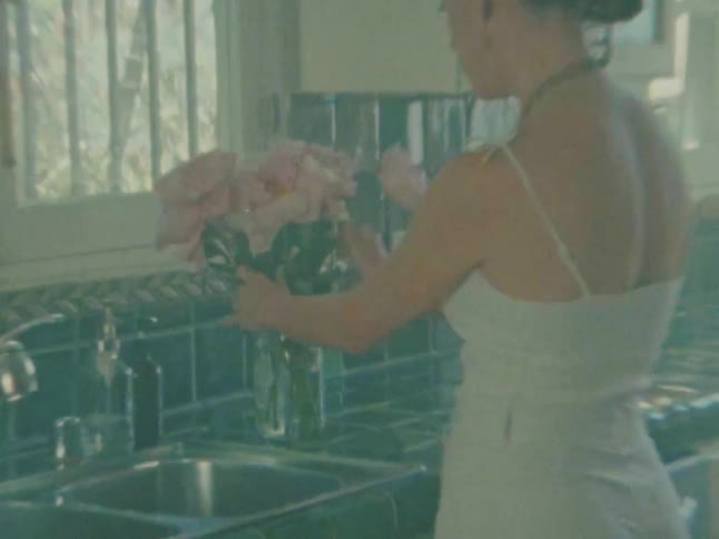 A person with hair tied up in a bun wearing a white dress stands facing a kitchen counter with a sink they are holding a large bouquet of pink roses and are in the process of placing them into a glass vase on the counter