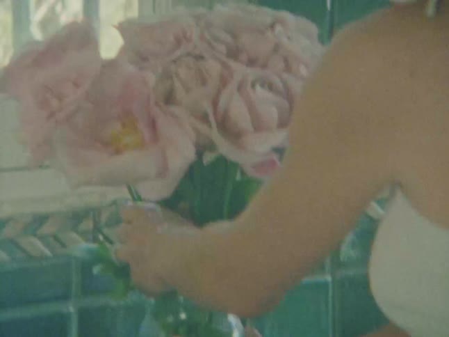 A person in a white top places a large bouquet of pink roses into a glass vase the setting appears to be a kitchen or bathroom with tiled walls visible behind the person and the flowers
