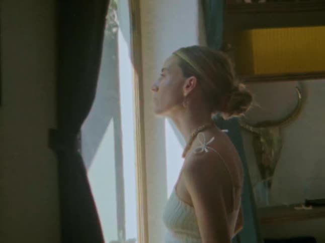 A woman wearing a lightcolored possibly white top is shown from the waist up she then turns to her right and looks out of a window which is framed by dark curtains