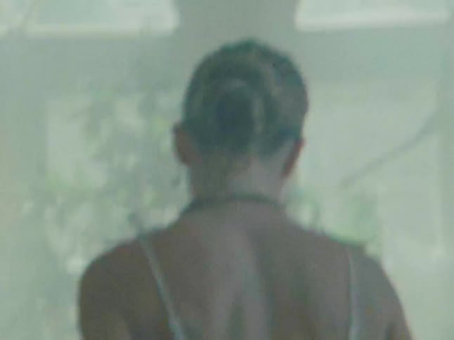 A woman with her hair in a bun enters a softly lit room with potted plants against a hazy background