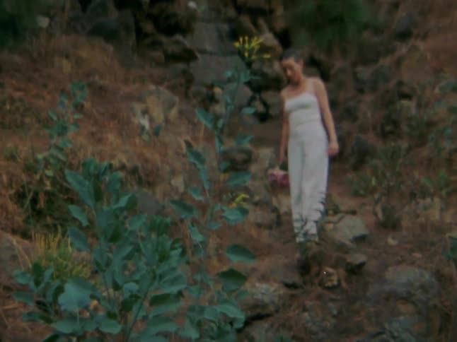 Woman standing on a rocky earthy path surrounded by sparse vegetation she is wearing lightcolored clothes and is holding a bouquet