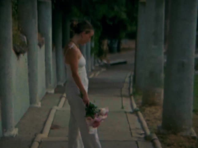 A person in white pants and a tank top walks along a stone pathway holding a bouquet of pink flowers eventually turning and walking away from the camera past several pillars