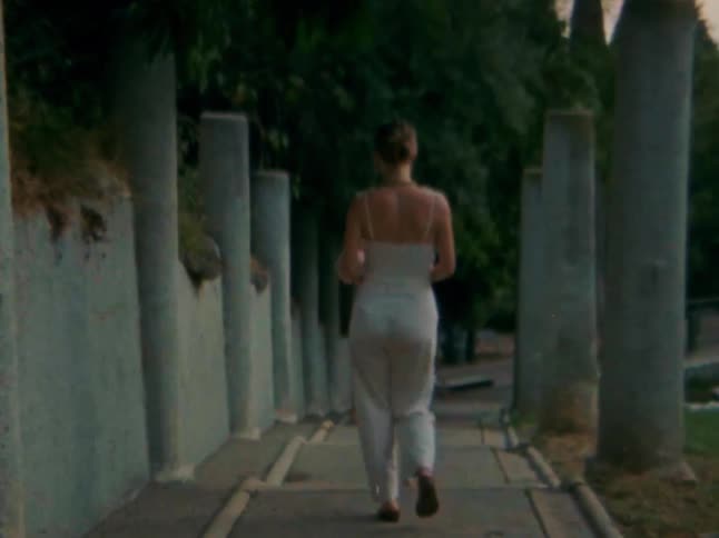 A woman walking down a path flanked by a series of stone columns on both sides and shes wearing lightcolored jumpsuit with a bouquet in her hand
