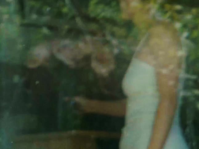 A woman in a white dress is seen through a window with green foliage reflected or visible behind her she is holding a bunch of flowers