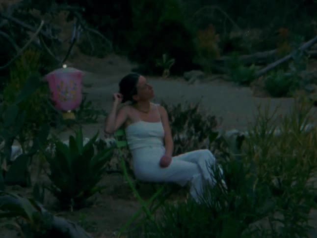 A woman in a lightcolored outfit is sitting on a chair in a natural somewhat unkempt outdoor setting surrounded by various plants and bushes to her left a pink decorative lantern hangs near some foliage