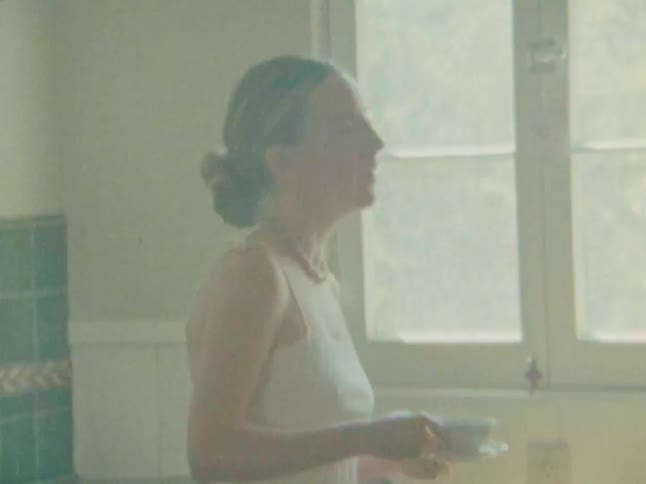 A woman in a lightcolored top stands in a kitchen with green tiled countertops she picks up a mug from the counter brings it to her mouth and takes a drink