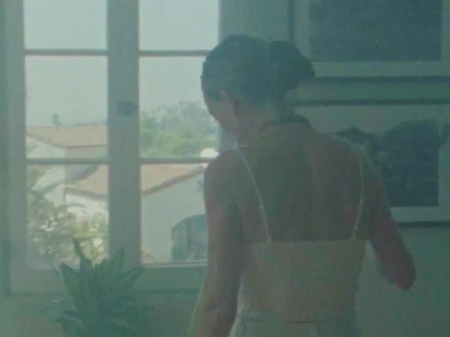 A woman holding a glass of water stands near a window then kneels down to water several potted plants she pours water from the glass into the soil of the plants