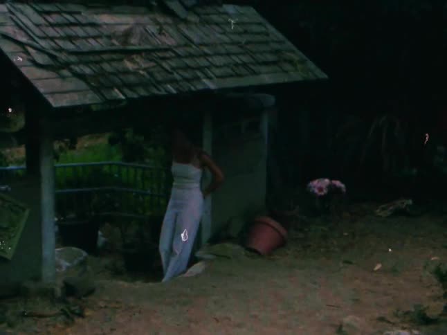 Person in white clothing leaning against a small rustic structure outdoors surrounded by pots and vegetation