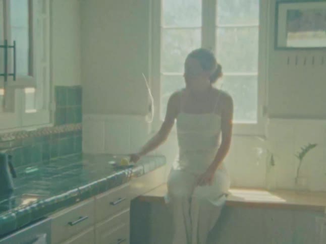 A woman in a lightcolored top is standing in a kitchen holding a yellow fruit possibly a lemon and appears to be wiping something on the counter she then sits down on the counter by a sunlit window