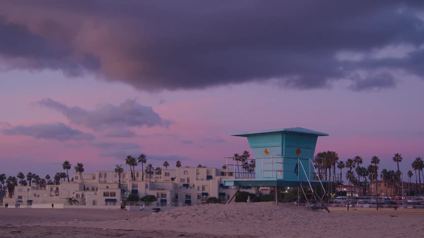 The video displays a picturesque beach scene at what appears to be either sunrise or sunset with a vibrant sky showing hues of pink and purple a prominent turquoise lifeguard tower stands on the sandy beach with white buildings and palm trees silhouett