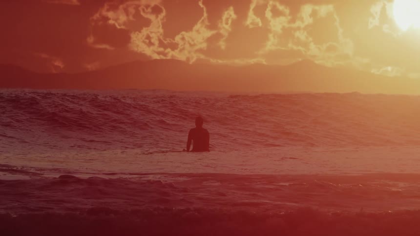 A silhouetted figure of a person sits on a surfboard in the ocean with waves surrounding them as the sun sets in an orange and yellow sky with clouds