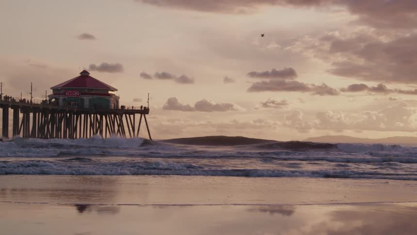 The video shows a pier extending into the ocean with a building at its end under a cloudy sky waves are breaking on the sandy beach in the foreground with distant mountains visible on the horizon