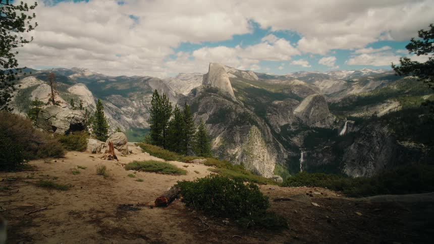 The video displays a breathtaking panoramic view of a mountainous landscape under a cloudy sky in the center a prominent rock formation likely half dome stands tall amidst pine trees with a waterfall visible in the distance on the right
