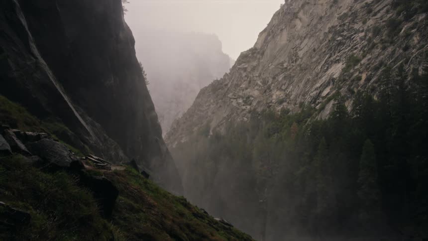 The video displays a narrow deep gorge with towering rocky cliffs on both sides disappearing into a thick mist or fog in the distance the sides of the gorge are sparsely covered with green vegetation especially in the foreground